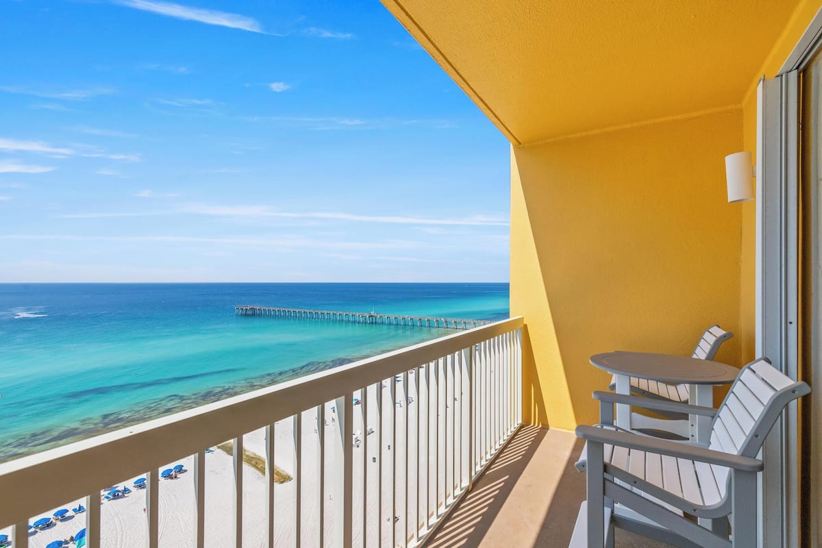 A balcony with a view of the ocean and a pier. There are two chairs and a small table.