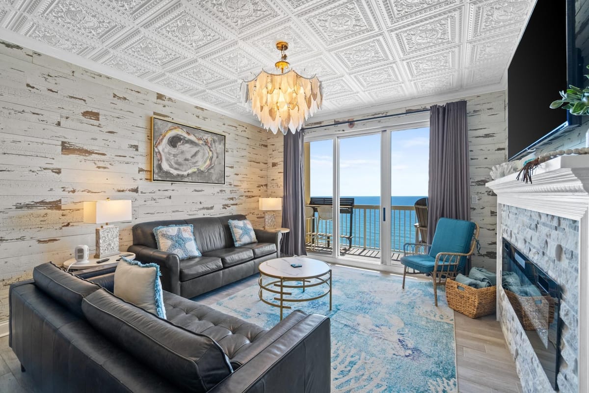A spacious living room with a large sofa, a coffee table, and a fireplace. There is a balcony with a view of the sea.
