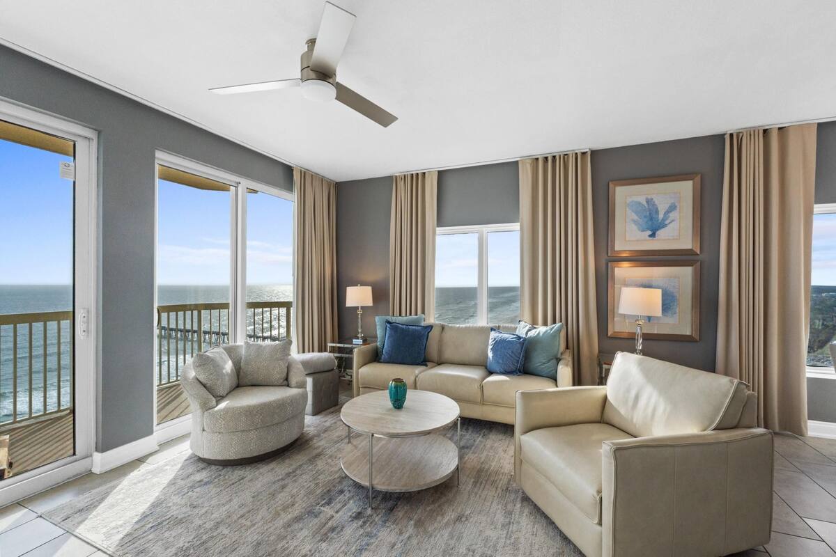 A spacious living room with a large window overlooking the ocean, a ceiling fan, and a view of the sea.