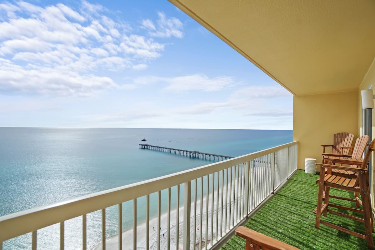 A balcony with a view of the ocean, a pier in the distance, and wooden chairs.