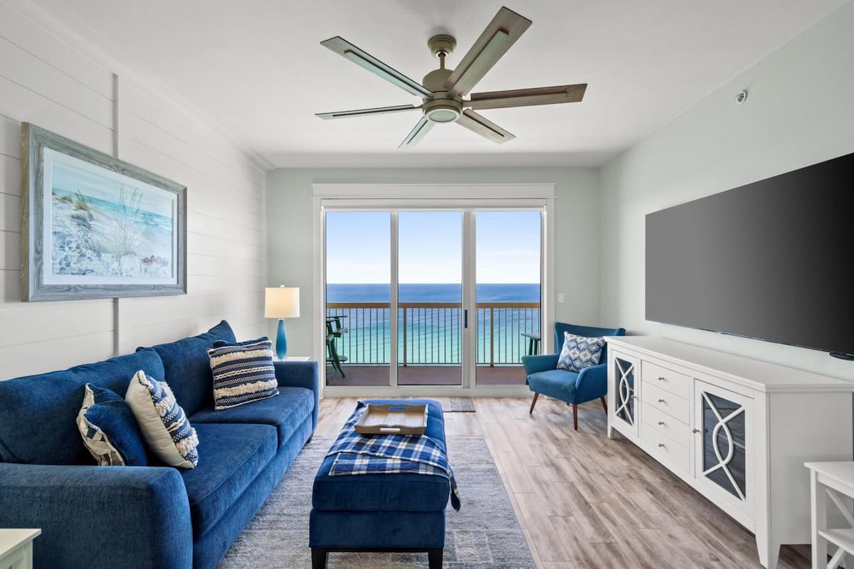 A living room with a blue sofa, a coffee table, a ceiling fan, a large TV, and a balcony with a view of the ocean.