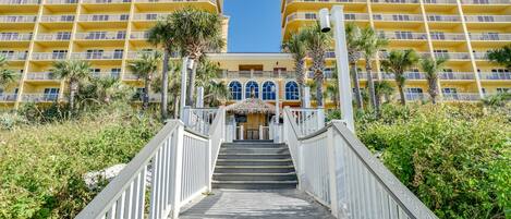 A staircase leading to a yellow building with balconies, surrounded by greenery and palm trees.