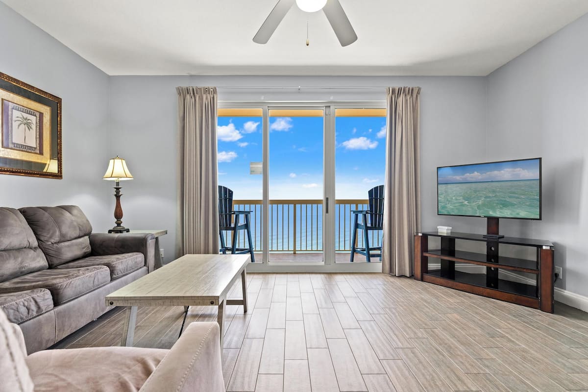 A living room with a sofa, a coffee table, a TV on a stand, and a balcony with a view.
