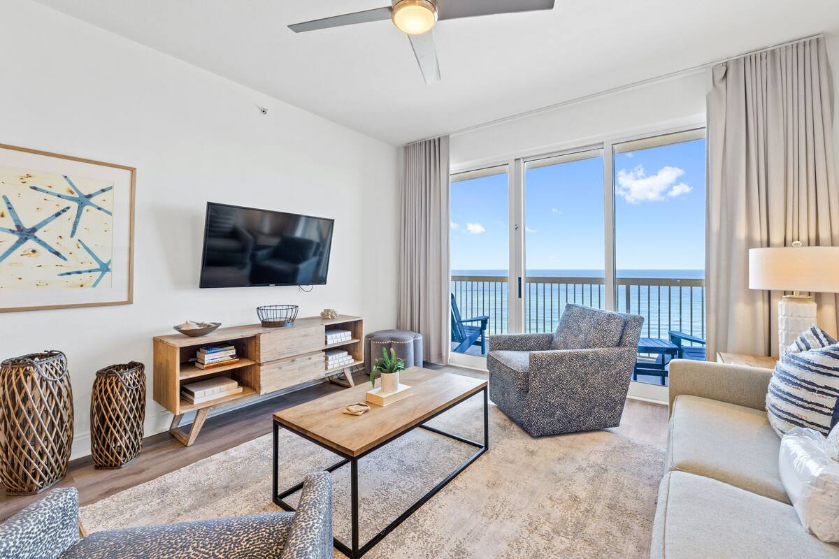 A modern living room with a flat-screen TV, wooden furniture, a large window with a view of the sea, and a ceiling fan.