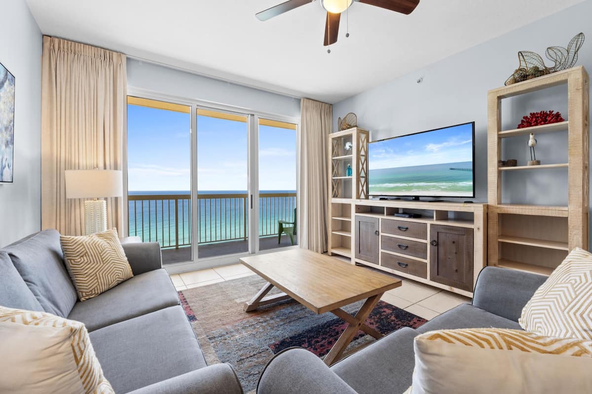 A modern living room with a large TV, a sofa, a coffee table, and a view of the ocean through large sliding doors.