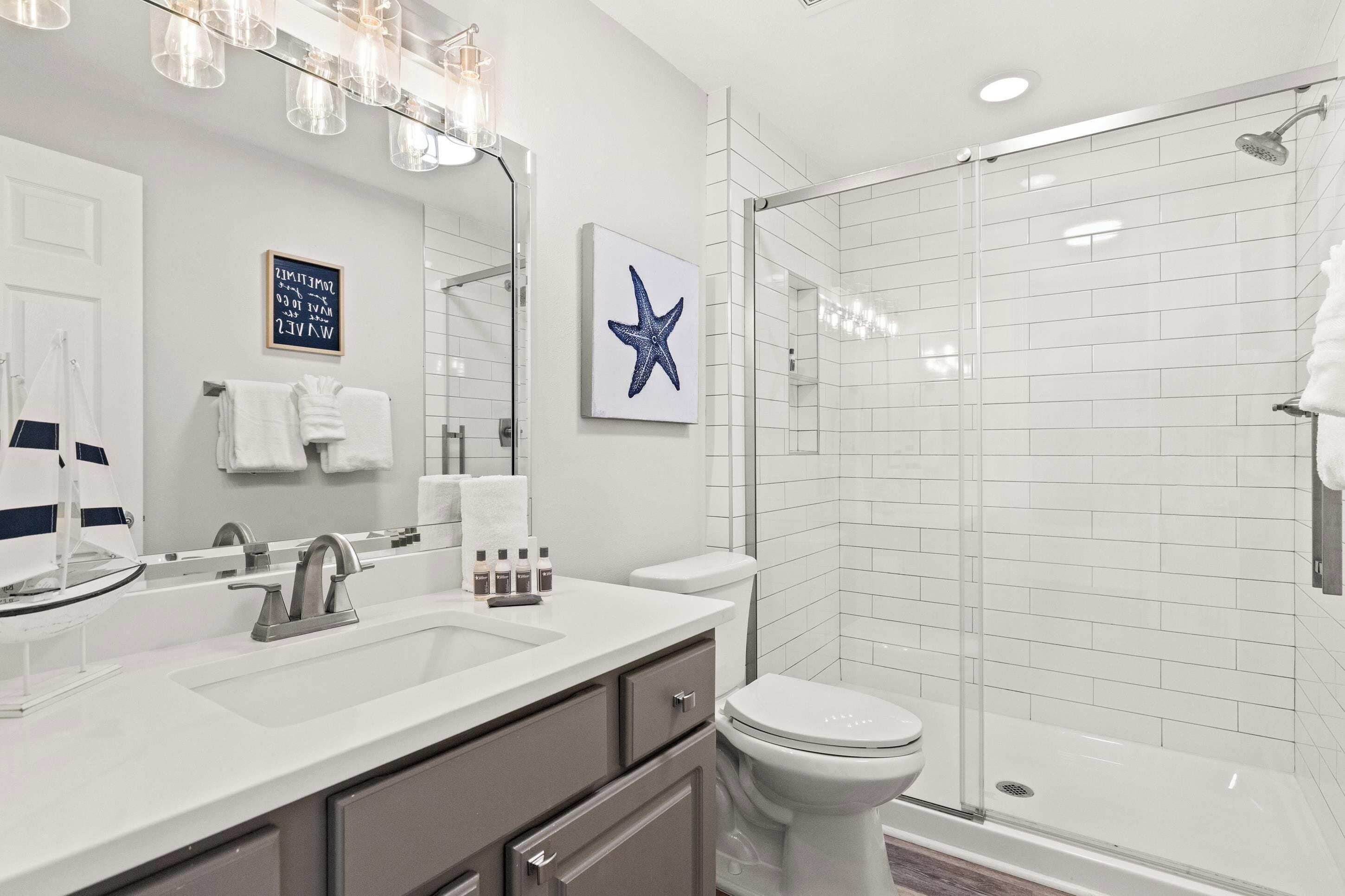 A modern bathroom with a white sink, mirror, and toilet, a glass shower enclosure, and a starfish decoration on the wall.