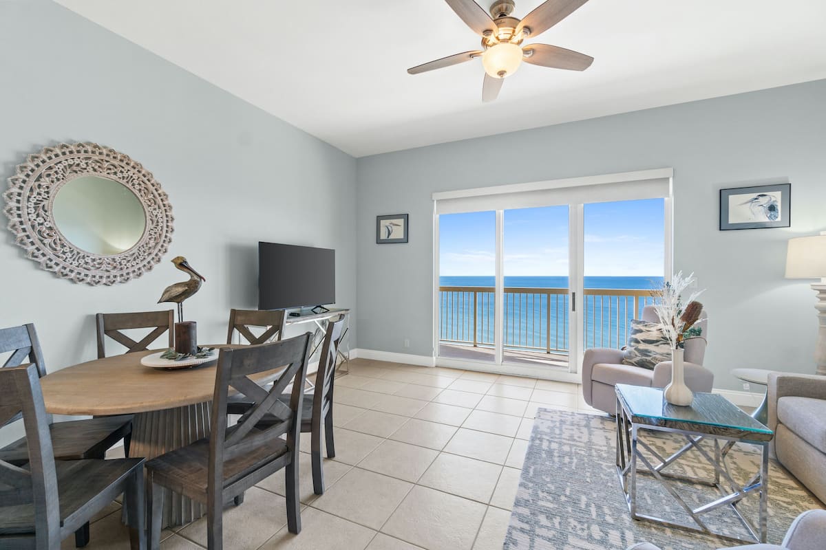 A spacious living area with a dining table, chairs, a ceiling fan, a large window with a view of the ocean, and a balcony.