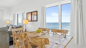 Una sala de estar con una mesa de comedor de madera, sillas, un sofá y vistas al océano a través de amplios ventanales.