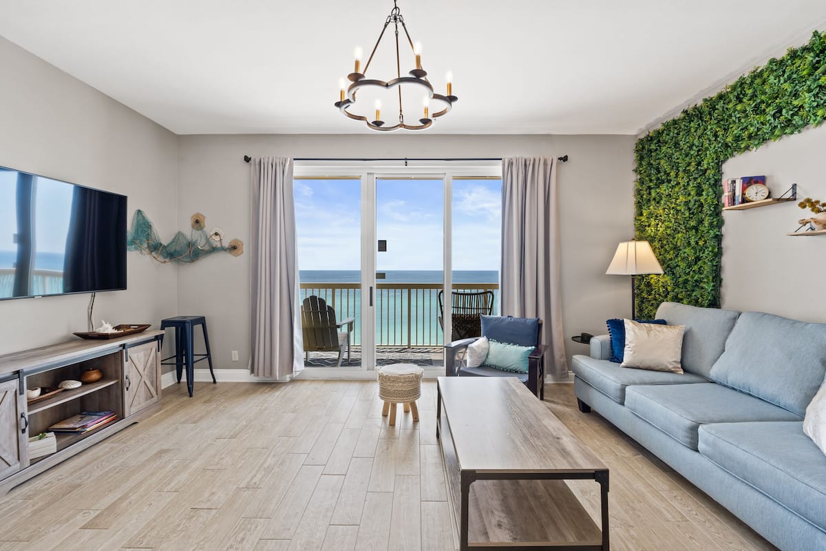 A living room with a sofa, a coffee table, a TV, a bookshelf, and a view of the ocean through large sliding doors.