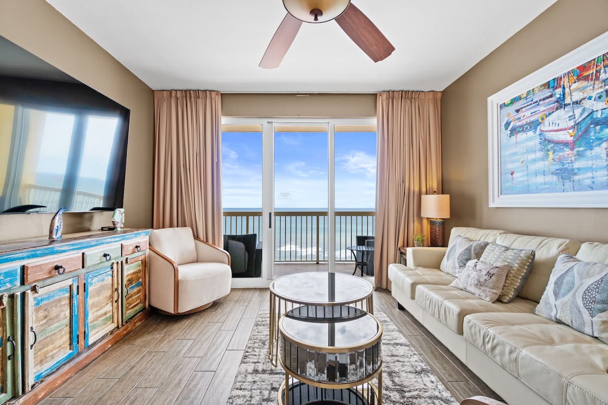 A spacious living room with a large window, a ceiling fan, a sofa, a coffee table, and a TV.