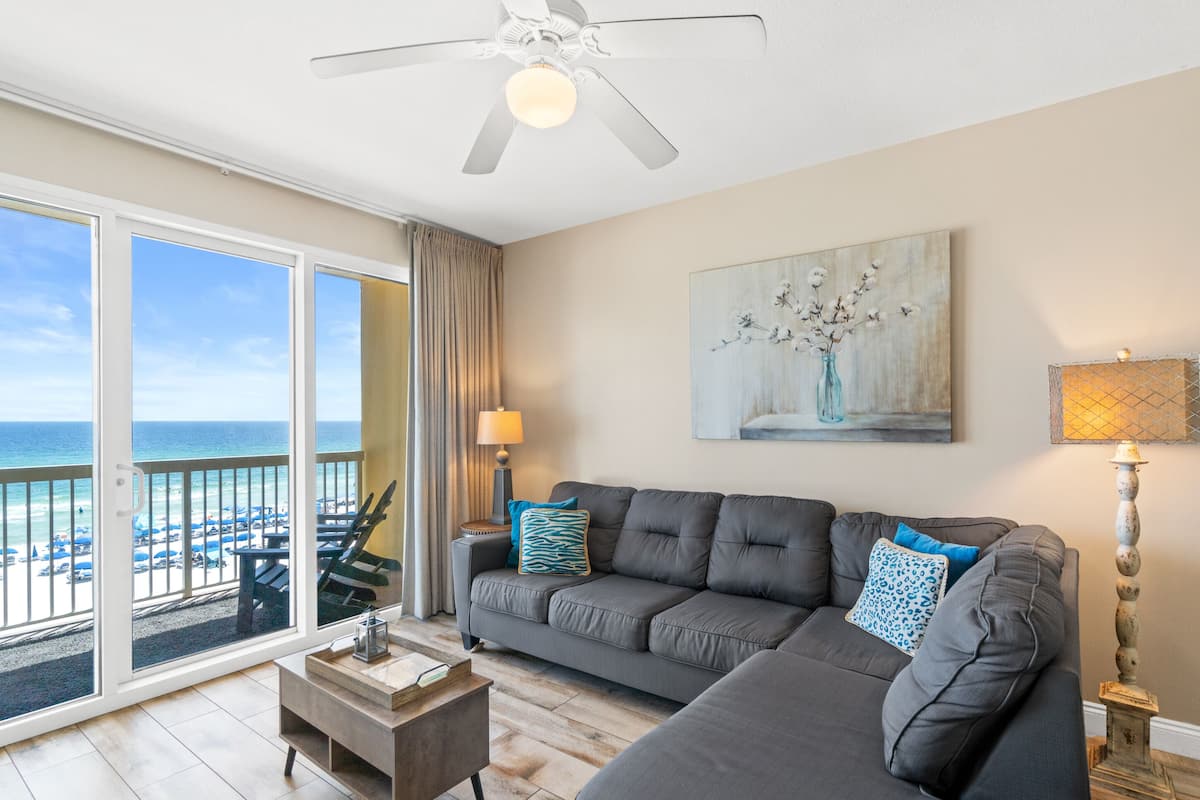 A living room with a gray sofa, a coffee table, a lamp, and a painting. There is a view of the beach through large windows.