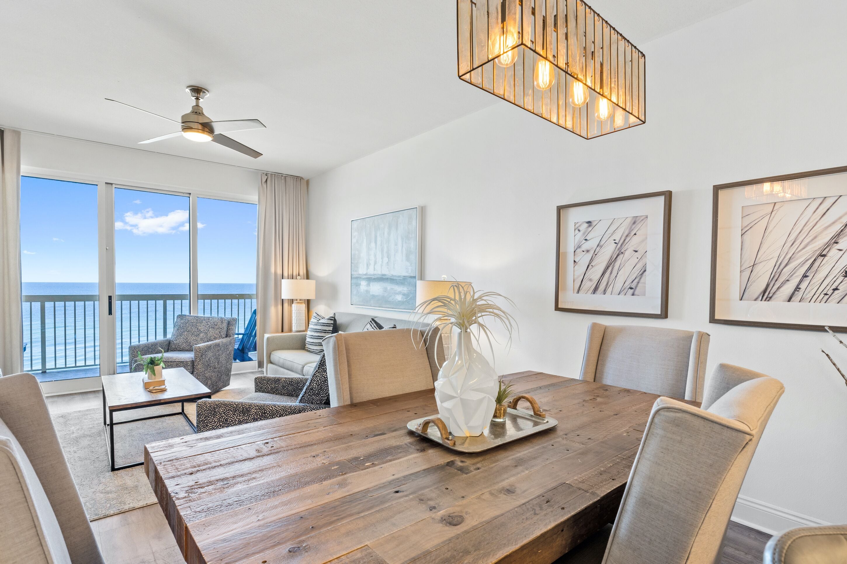 A spacious room with a wooden dining table, comfortable seating, a ceiling fan, and large windows offering a view of the ocean.