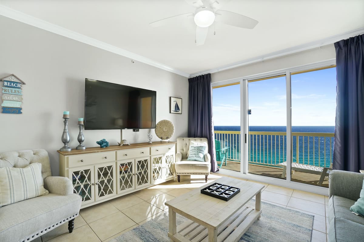A living room with a TV, a sofa, a coffee table, and a view of the ocean through large sliding doors.