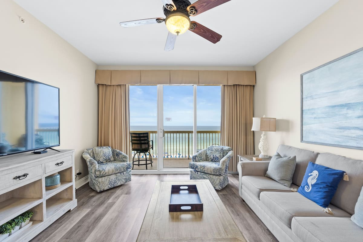A spacious living room with a flat-screen TV, two patterned armchairs, a beige sofa, and a ceiling fan.
