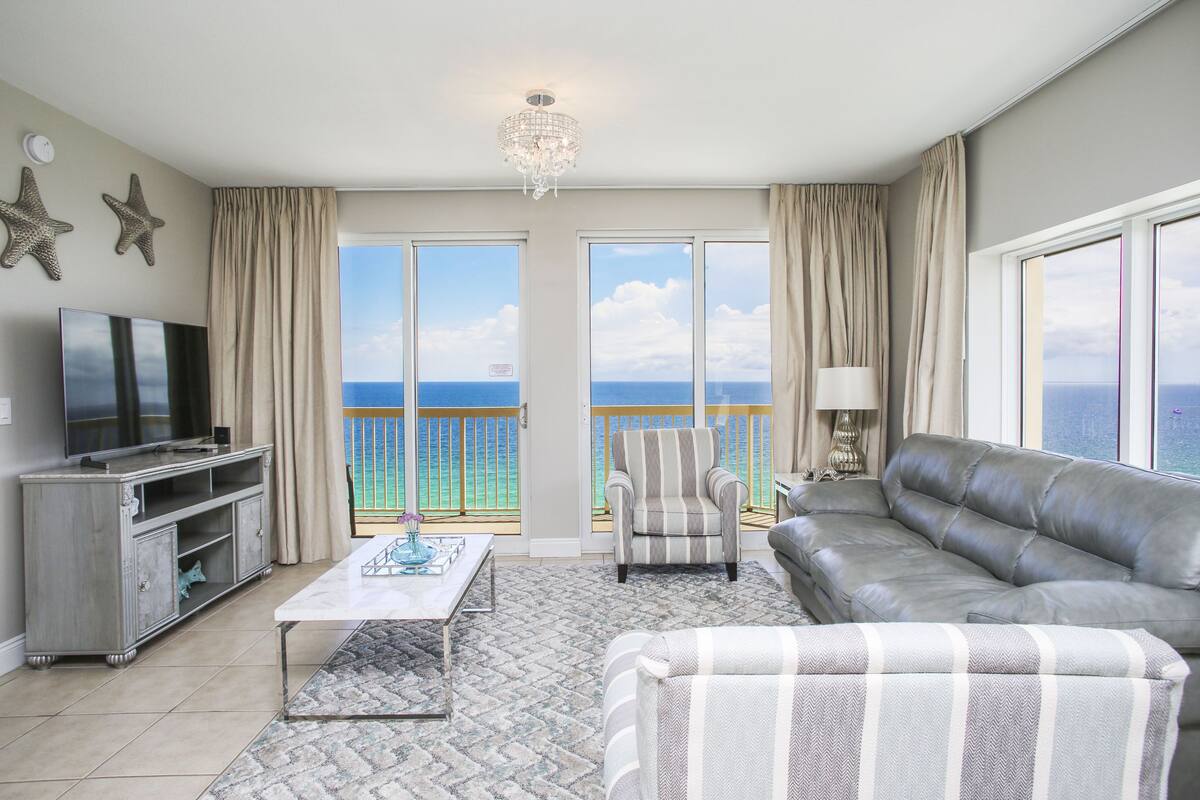 A living room with a television, sofa, armchair, and a view of the ocean through large windows.