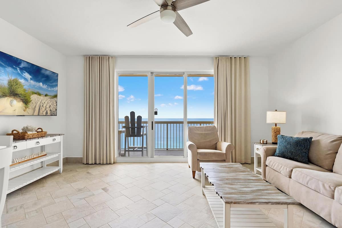 A spacious living room with a balcony, a ceiling fan, a sofa, a coffee table, and a TV on the wall.