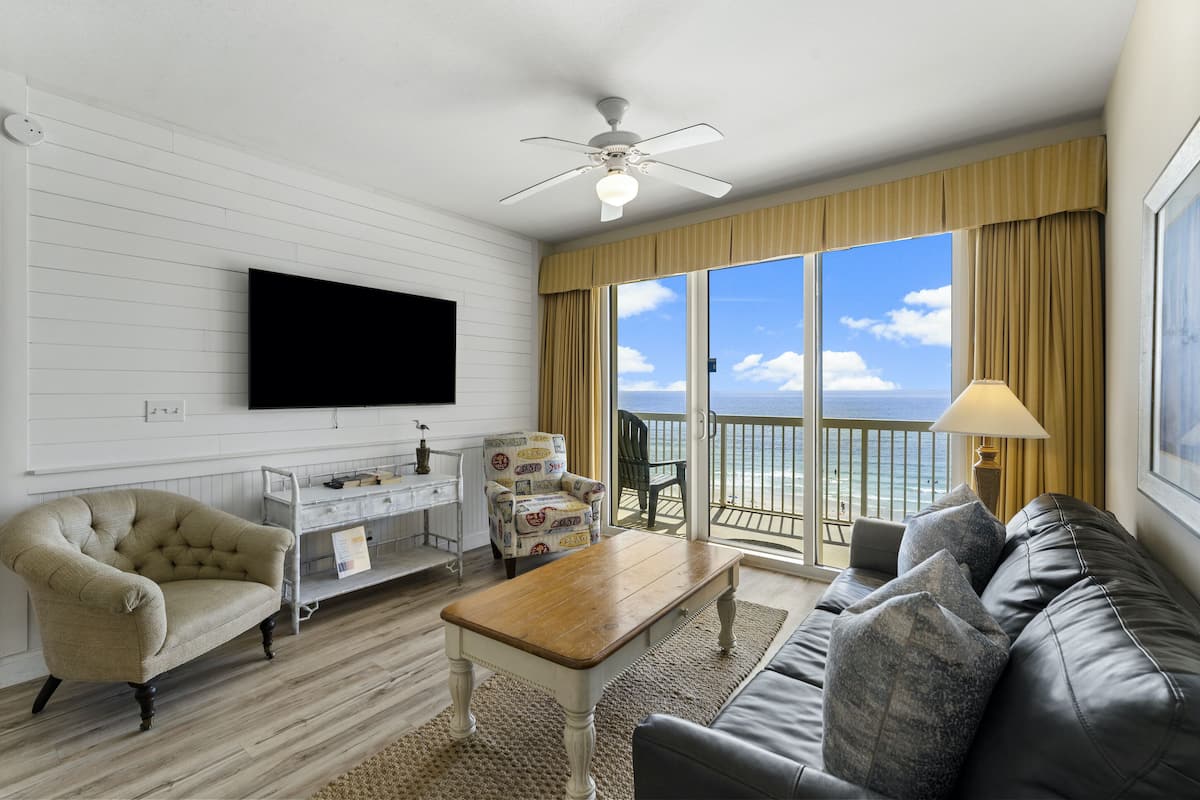A living room with a flat-screen TV, a beige armchair, a wooden coffee table, a leather sofa, and a view of the ocean through large windows.