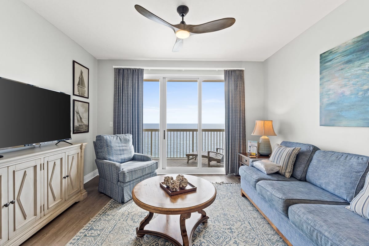 A living room with a television, a sofa, a chair, a coffee table, and a balcony with a view of the ocean.