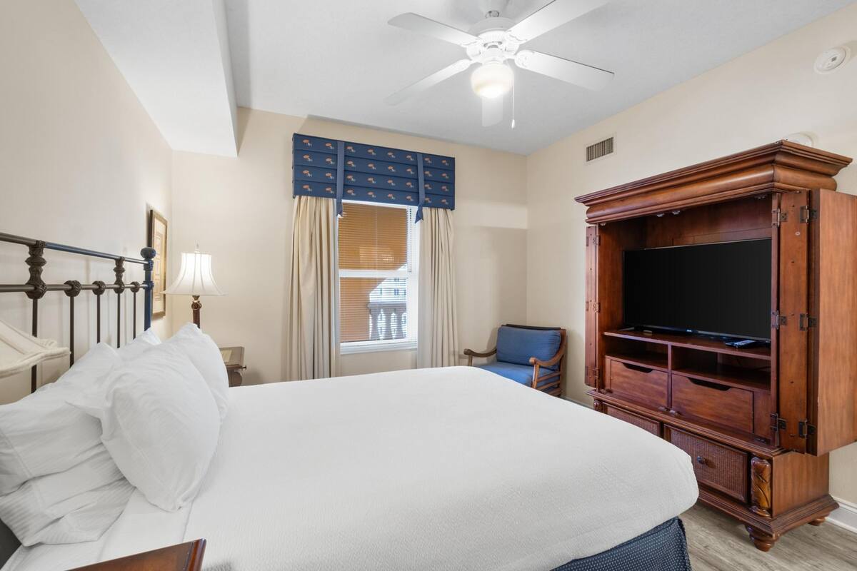 A hotel room with a bed, a television, a ceiling fan, a window with curtains, and a chair.
