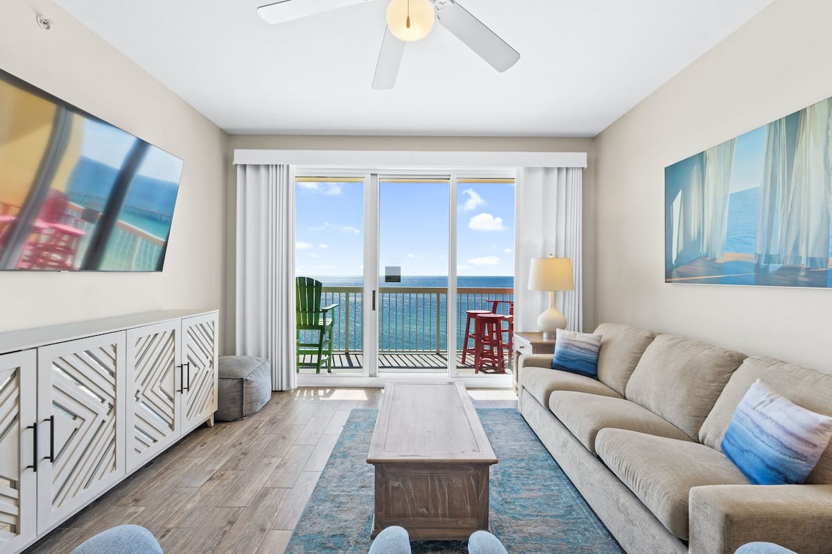 A spacious living room with a large window, a ceiling fan, a sofa, a coffee table, and a view of the ocean.