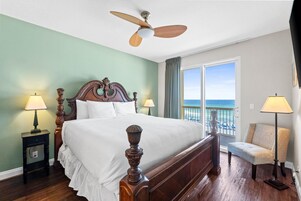 A hotel room with a large bed, a ceiling fan, a view of the ocean, and a chair by the window.