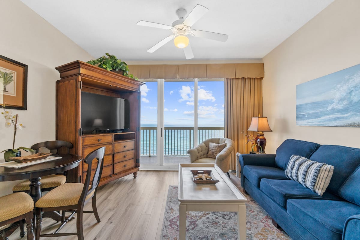 A living room with a TV, a blue sofa, a dining table, and a view of the ocean through large windows.