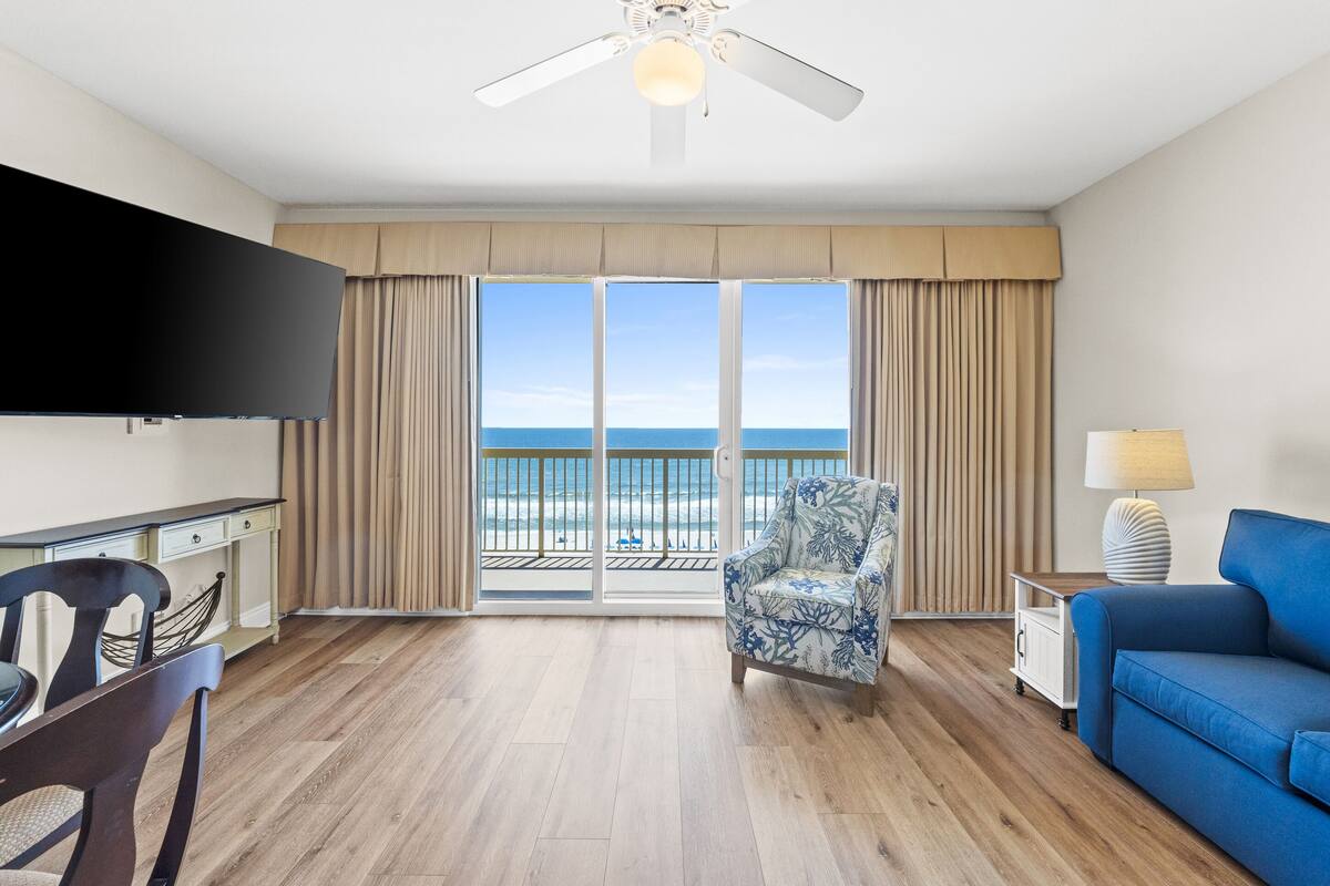 A hotel room with a wooden floor, a blue sofa, a patterned armchair, a flat-screen TV, a side table with a lamp, and a balcony with a view of the beach.