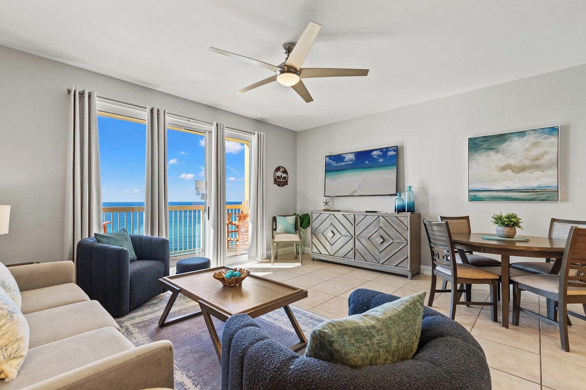 A spacious living room with a large window offering a view of the ocean, a ceiling fan, a dining area with a table and chairs, and a kitchen area with a cabinet and a plant.