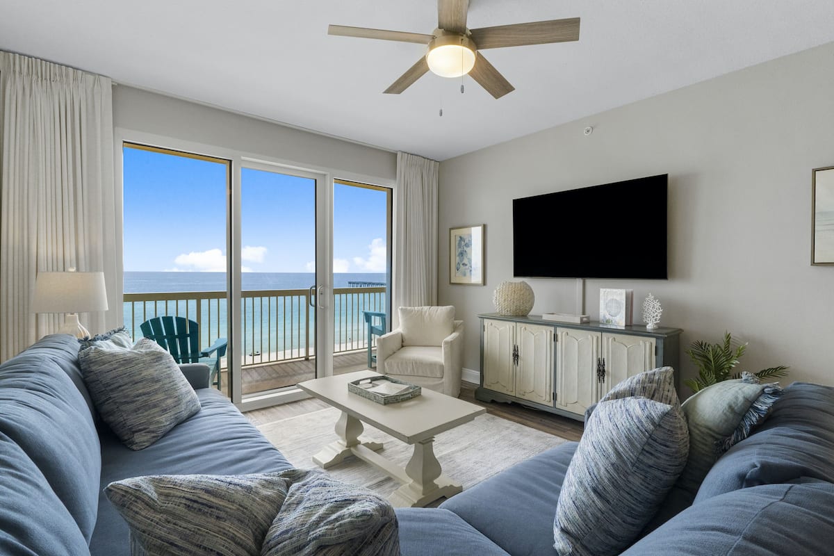 A living room with a large sofa, a coffee table, a television, and a view of the ocean.