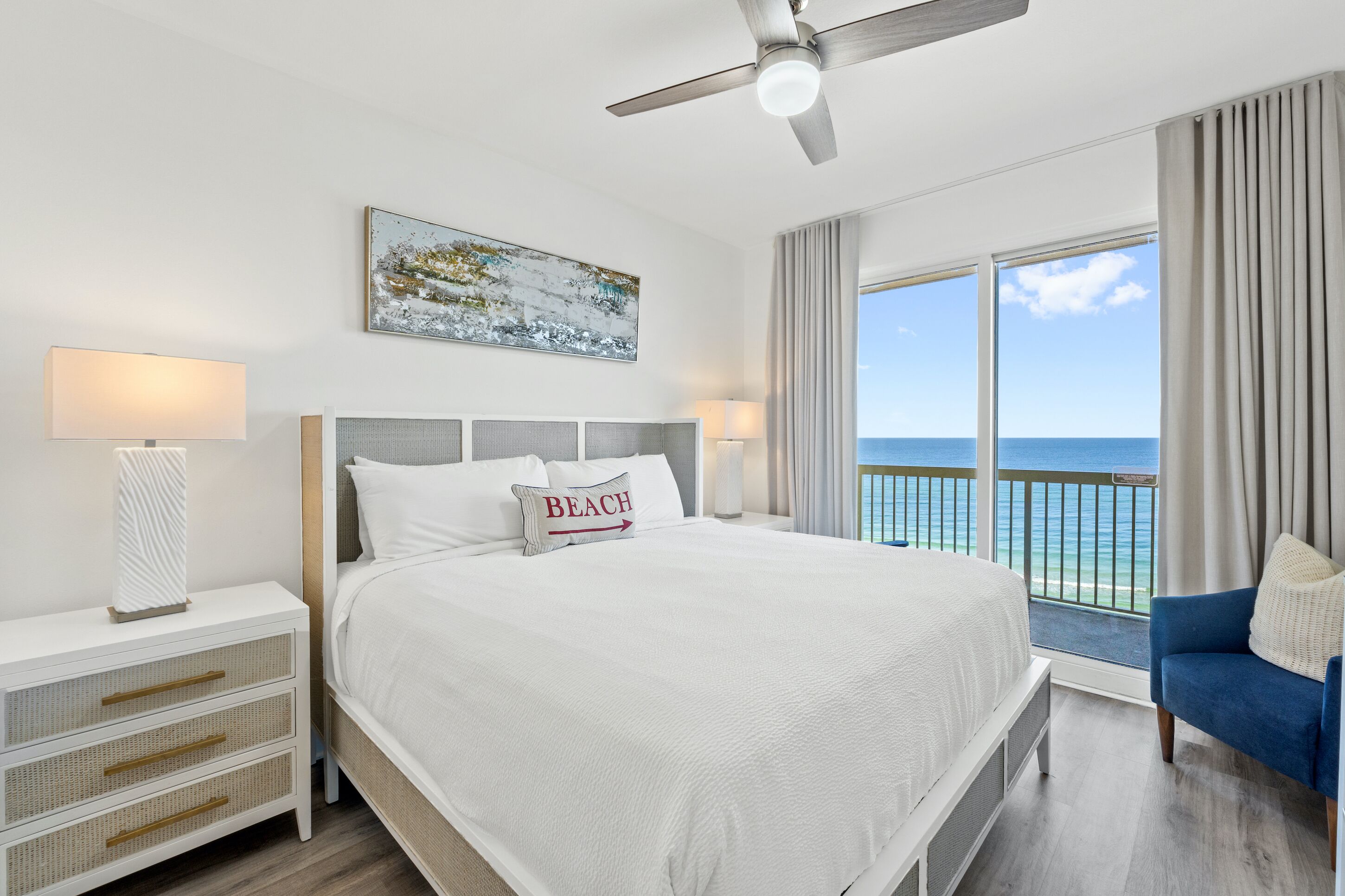 A hotel room with a large bed, a ceiling fan, a view of the ocean, and a balcony with curtains.