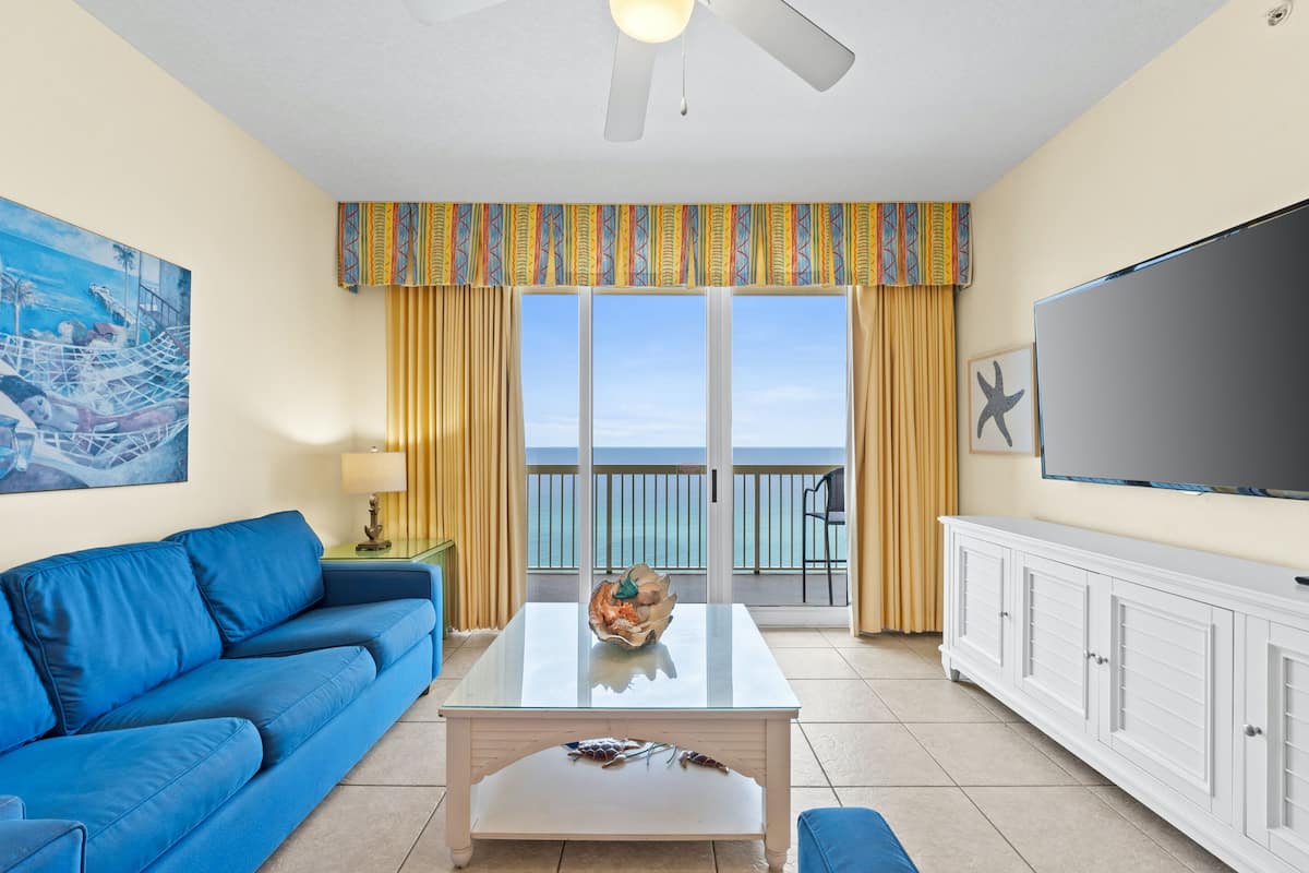 A living room with a blue sofa, a glass coffee table with decorative items, a flat-screen TV, and a balcony with a view of the ocean.