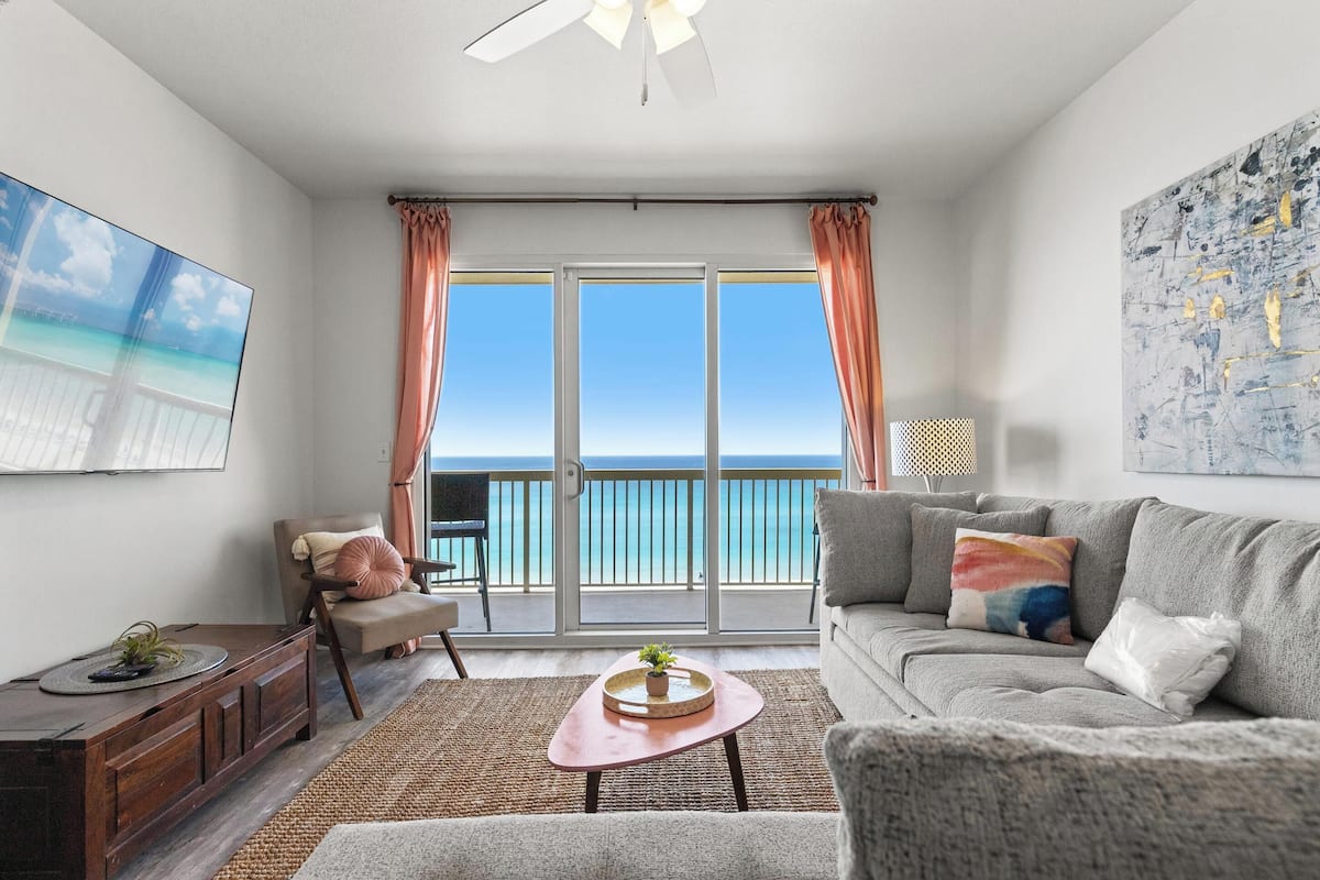A living room with a sofa, a coffee table, a chair, a TV, and a balcony with a view of the ocean.