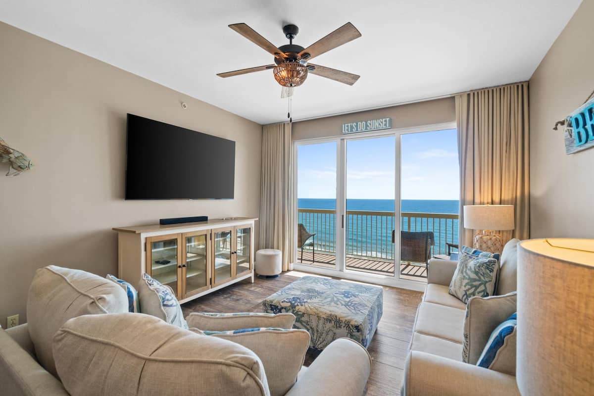 A living room with a flat-screen TV, a ceiling fan, a large window with a view of the ocean, and a sign that reads "LET'S DO SUNSET".