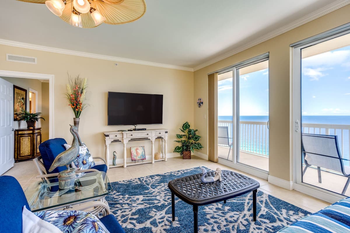 A living room with a large TV, a sofa, a coffee table, and a view of the ocean through large sliding doors.