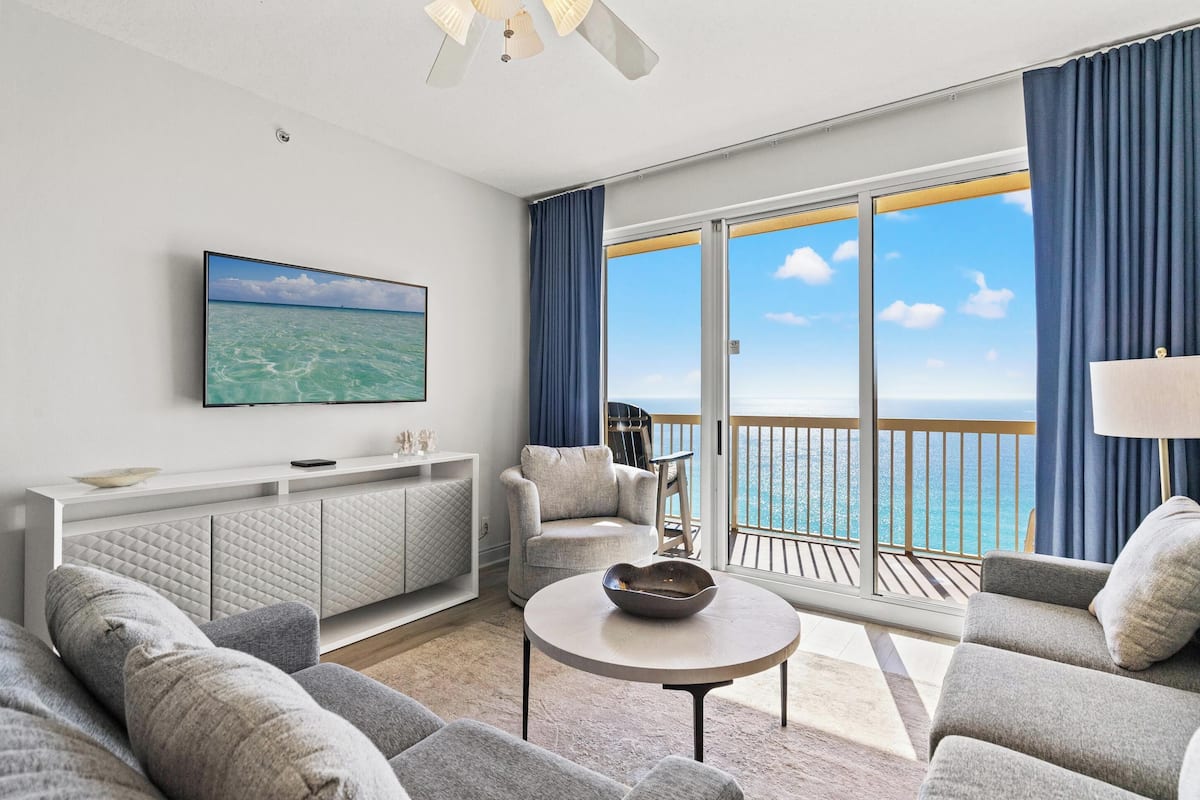 A modern living room with a large TV, comfortable seating, a balcony with a view of the ocean, and a white console table.