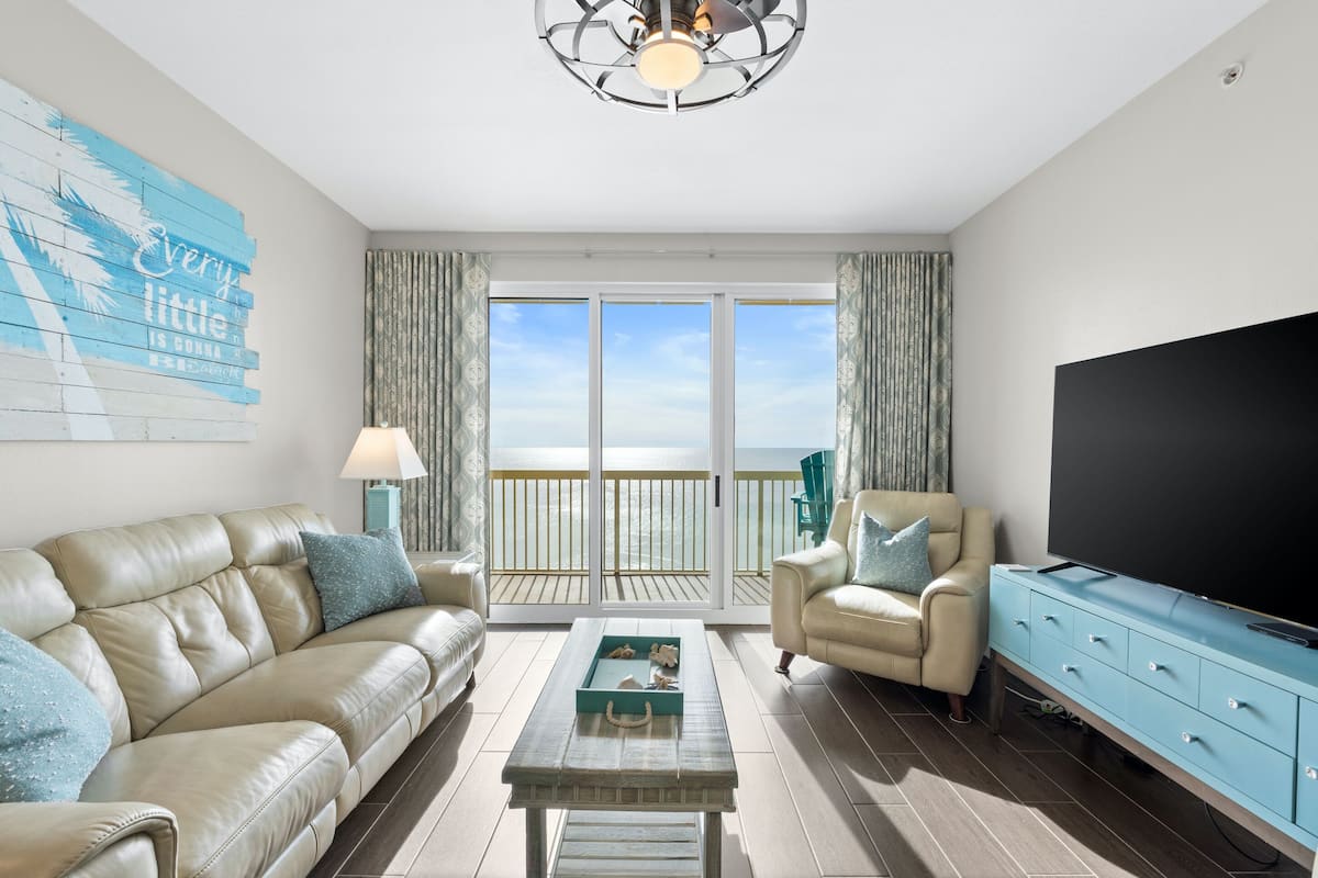 A modern living room with a leather sofa, a coffee table, a TV, and a balcony with a view of the ocean.