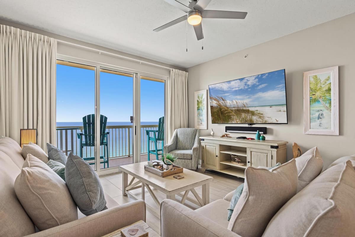 A living room with a TV, a sofa, a coffee table, and a balcony with chairs.