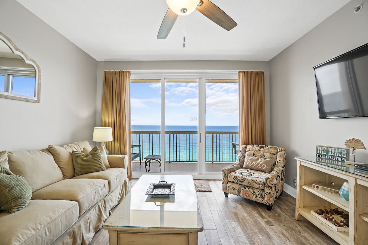 A living room with a sofa, armchair, coffee table, and a TV. It features a balcony with a view of the ocean.