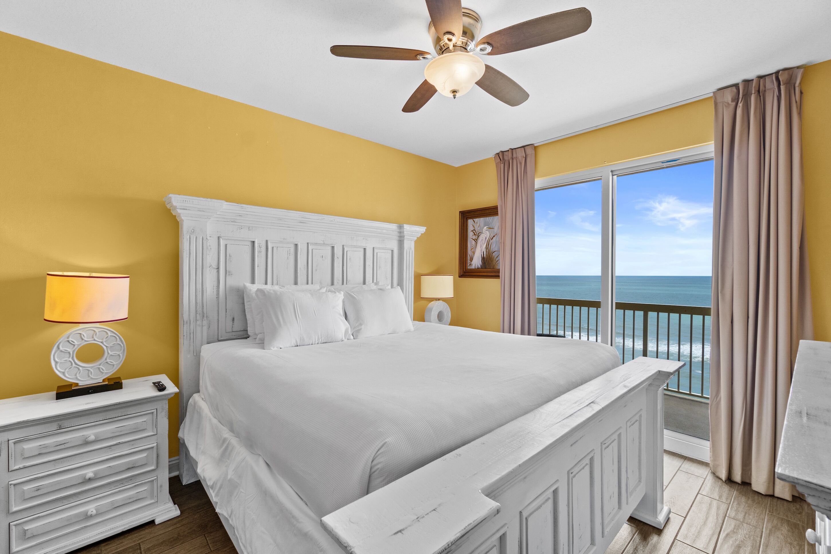 A bedroom with a white bed, a ceiling fan, a nightstand with a lamp, and a view of the ocean through large windows.