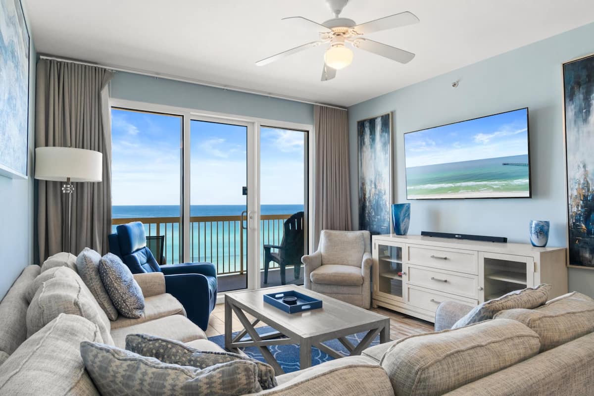 A spacious living room with a large sofa, a coffee table, and a flat-screen TV. There is a balcony with a view of the ocean.