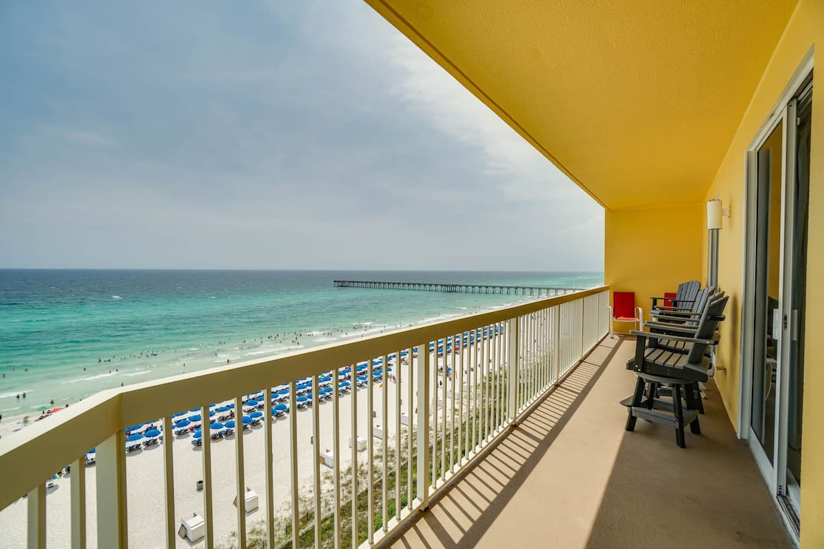A balcony with a view of the beach and a pier. There are beach chairs and umbrellas on the balcony.