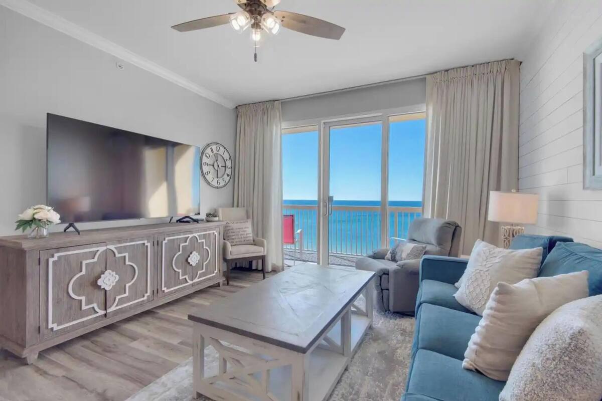 A living room with a TV, a coffee table, a sofa, a chair, and a balcony with a view.
