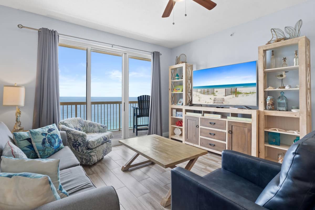 A spacious living room with a large window, a wooden coffee table, a sofa, a TV, and a wooden cabinet with decorative items.