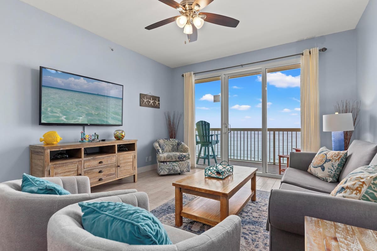 A living room with a TV, wooden furniture, a ceiling fan, and a view of the ocean through a sliding door.