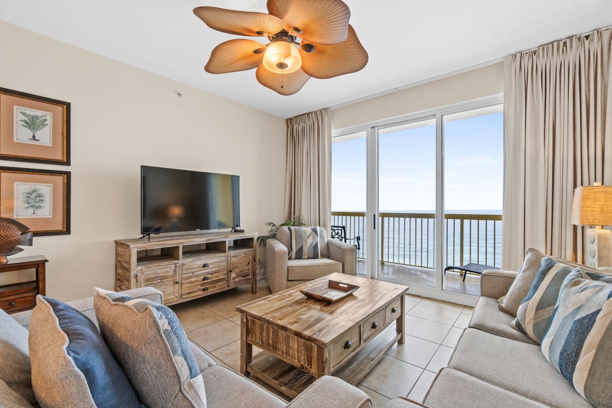 A spacious living room with a large flat-screen TV, a wooden coffee table, and a sofa. There is a balcony with a view of the sea.