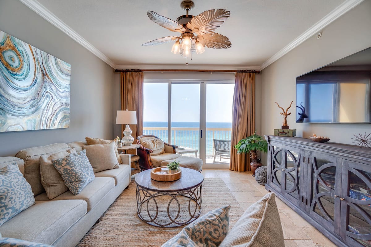 A spacious living room with a large sofa, a coffee table, and a ceiling fan. There is a view of the ocean through large windows.