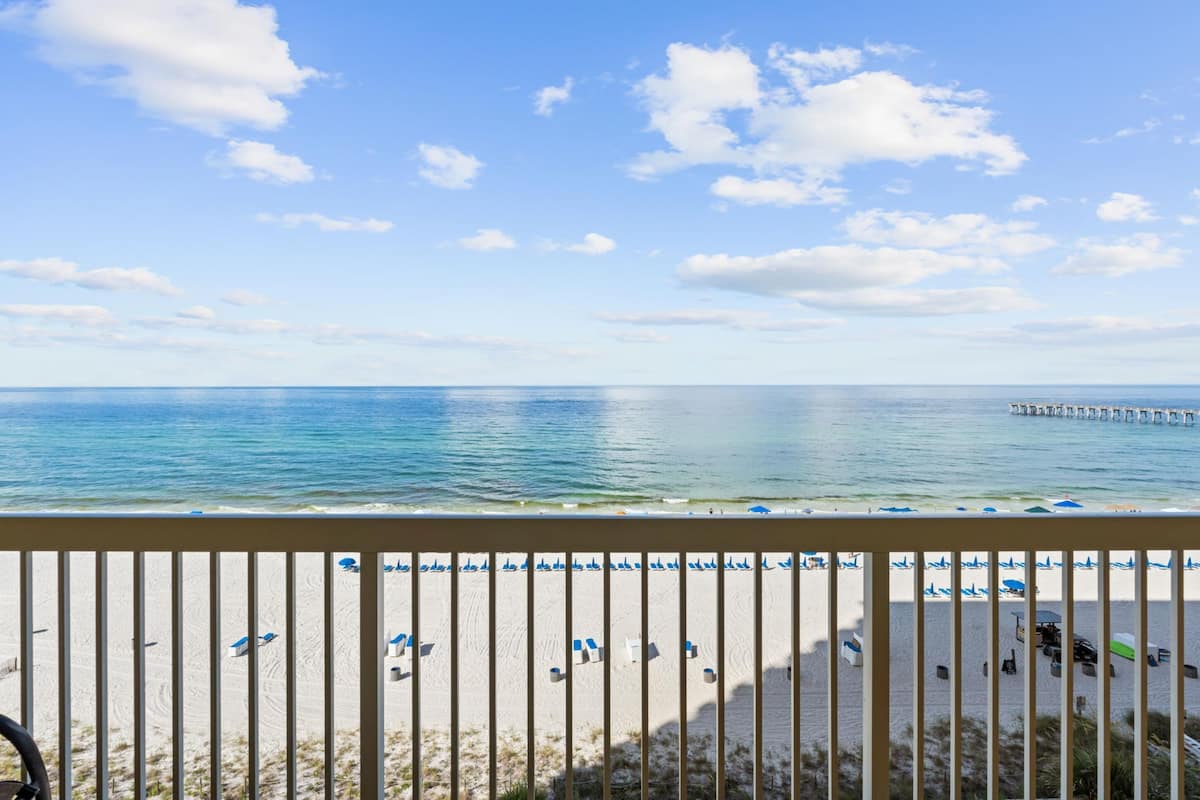 A beach view from a balcony with a railing, beach umbrellas, and a pier in the distance.
