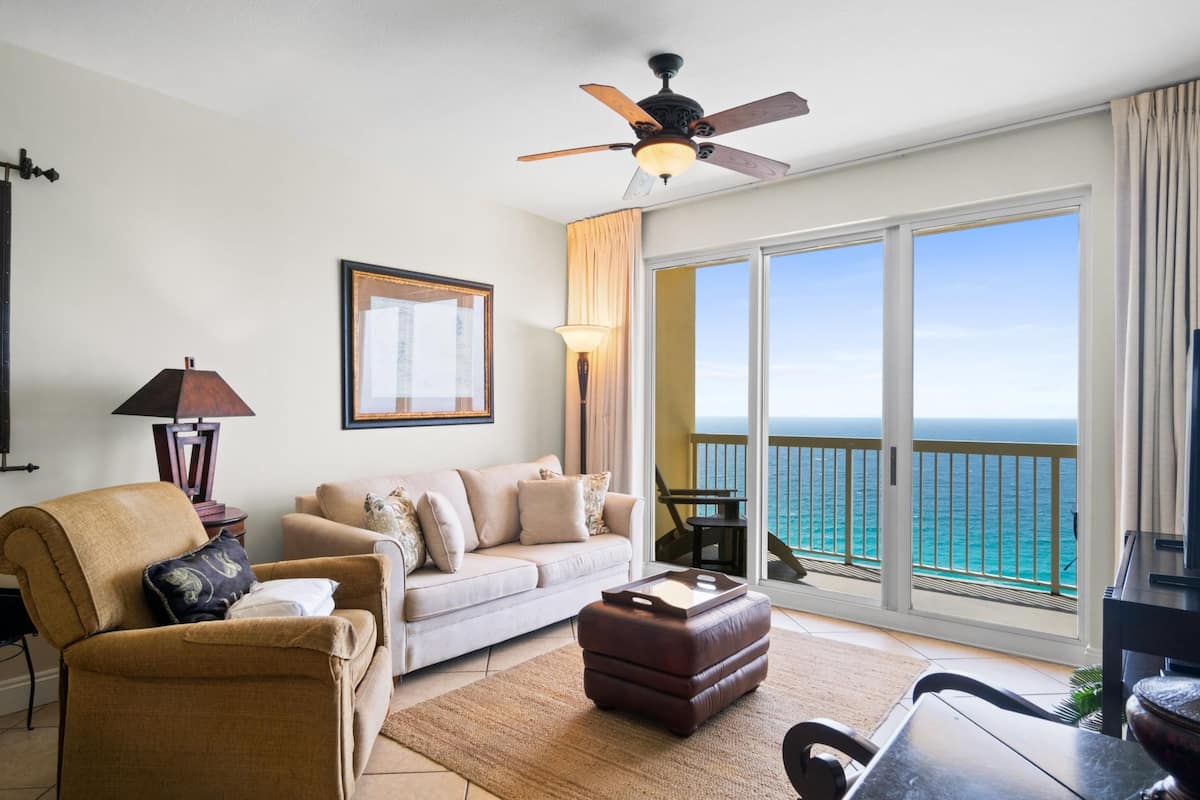 A living room with a sofa, armchair, and ottoman, a ceiling fan, a lamp, and a large window with a view of the ocean.