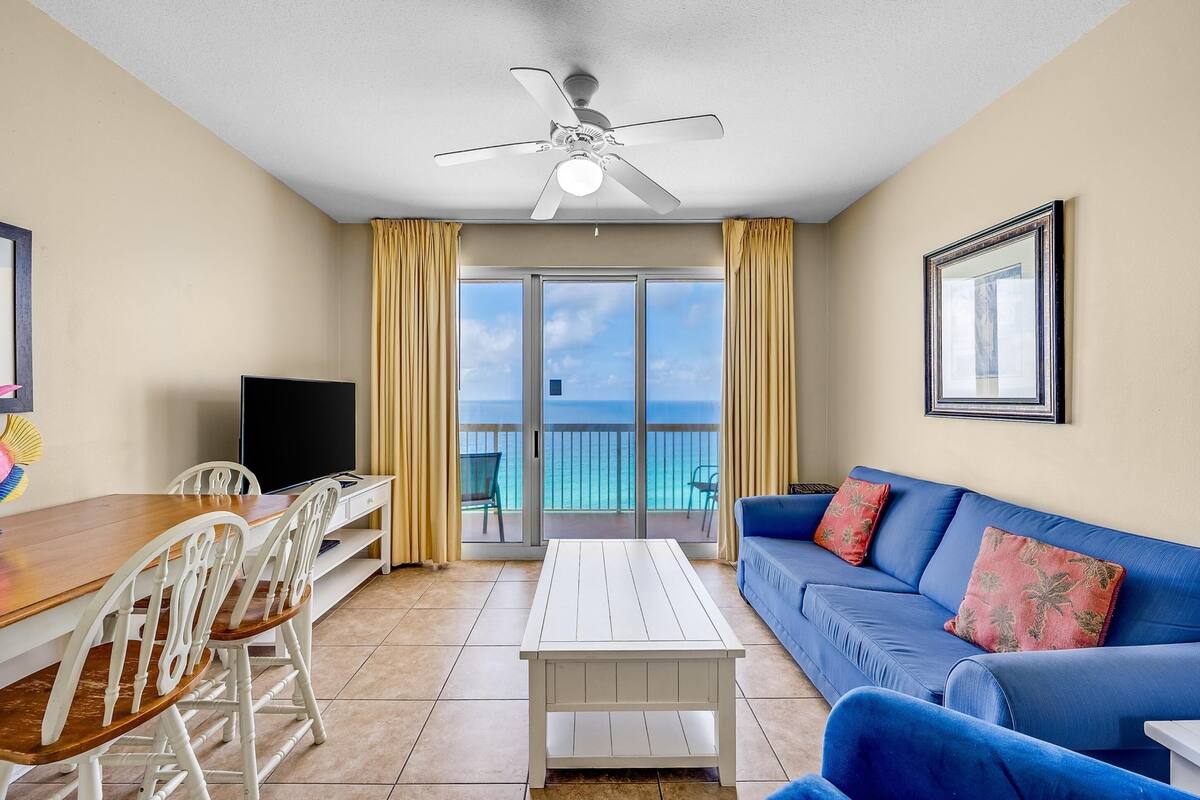 A room with a white ceiling fan, a TV, a wooden dining table with chairs, a blue sofa with decorative pillows, and a balcony with a view of the ocean.