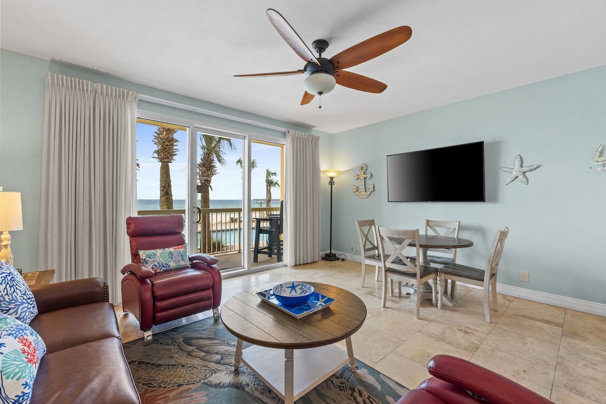 A living room with a ceiling fan, a TV mounted on the wall, a dining table with chairs, and a view of the ocean through large windows.